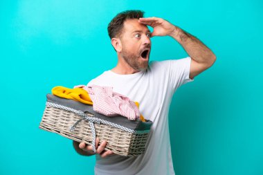 Orta yaşlı bir kadın mavi arka planda izole edilmiş bir çamaşır sepeti tutuyor. Yan tarafa bakarken sürpriz jest yapıyor.