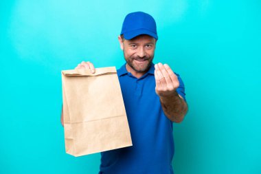 Orta yaşlı bir adam, mavi arka planda izole edilmiş bir paket yiyecek alıyor ve bizimle gelmesi için davet ediyor. Gelmene sevindim.