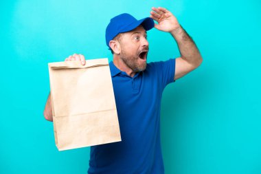 Orta yaşlı bir adam mavi arka planda izole edilmiş bir paket yiyecek alıyor. Yan tarafa bakarken sürpriz jest yapıyor.