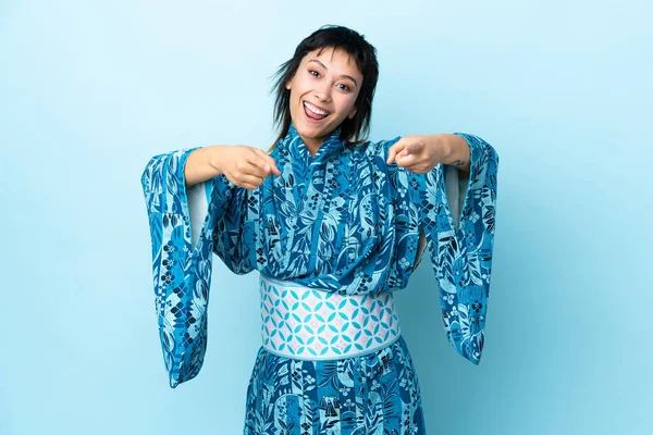 İzole edilmiş mavi arka planda kimono giyen genç bir kadın şaşırmış ve ön tarafı işaret ediyor.