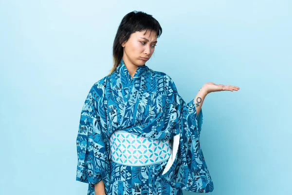 İzole edilmiş mavi arka plan üzerinde kimono giyen genç bir kadın. Kopyalama alanını şüpheyle tutuyor.