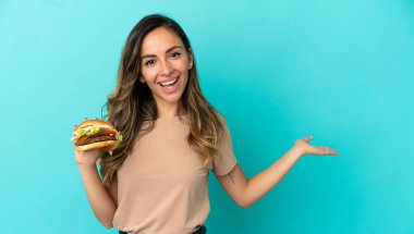 Soyutlanmış arka planda hamburger tutan genç bir kadın. Davet etmek için ellerini yana uzatıyor.