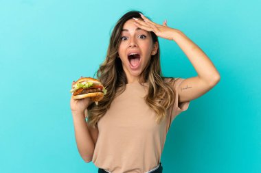 Arka planda hamburger tutan genç bir kadın. Yan tarafa bakarken sürpriz jest yapıyor.