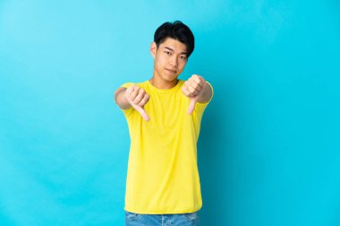 Mavi arka planda izole edilmiş genç bir Çinli iki eliyle baş parmağını aşağı gösteriyor.