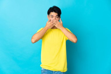 Mavi arka planda izole edilmiş genç Çinli adam elleriyle ağzını kapatıyor.