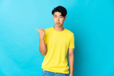 Genç Çinli adam mavi arka planda izole edilmiş mutsuz ve kenarı işaret ediyor.