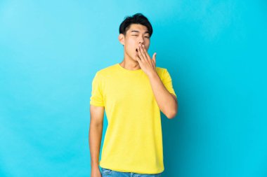 Genç Çinli bir adam mavi arka planda izole edilmiş esniyor ve elleriyle ağzını kapatıyor.