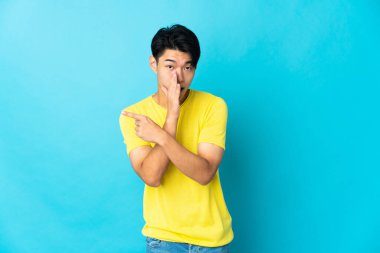 Mavi arka planda izole edilmiş genç Çinli bir adam bir ürün sunmak ve bir şeyler fısıldamak için kenarı işaret ediyor.