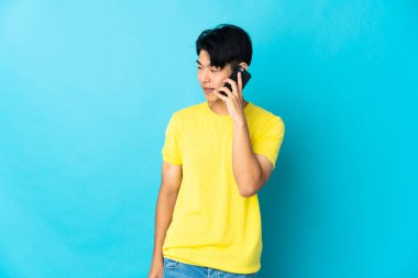 Genç Çinli bir adam mavi arka planda izole edildi. Cep telefonuyla biriyle sohbet ediyor.
