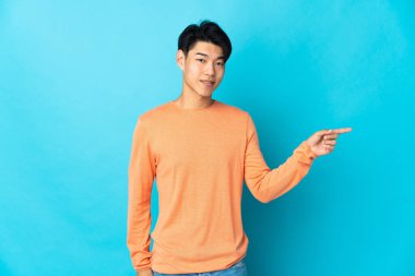 Genç Çinli adam mavi arka planda izole edilmiş, parmağını yana doğru gösteriyor.
