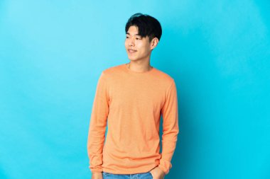 Genç Çinli adam mavi arka planda izole edilmiş, yan tarafa bakıyor ve gülümsüyor.