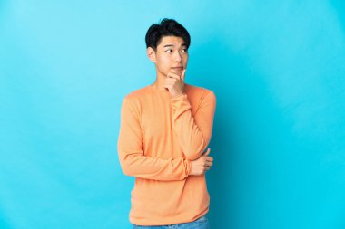 Genç Çinli bir adam mavi arka planda izole edilmiş, yukarı bakarken bir fikir düşünüyor.
