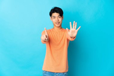 Mavi arka planda izole edilmiş genç Çinli adam parmaklarıyla altısını sayıyor.