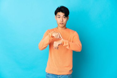 Genç Çinli adam mavi arka planda izole edilmiş, geç kalma jesti yapıyor.