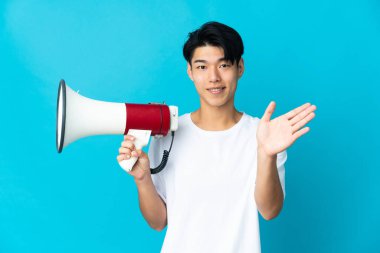 Mavi arka planda izole edilmiş genç bir Çinli megafon tutuyor ve mutlu bir ifadeyle selamlıyor.
