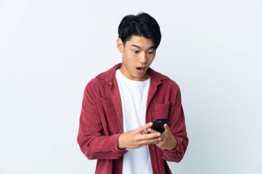 Genç Çinli adam beyaz arka planda izole edilmiş kameraya bakarken şaşırmış bir ifadeyle cep telefonunu kullanıyor.