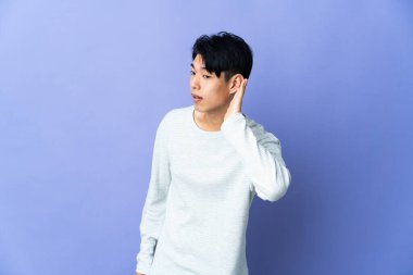 Mor arka planda izole edilmiş genç Çinli bir adam bir şeyler dinlerken elini kulağına koyar.