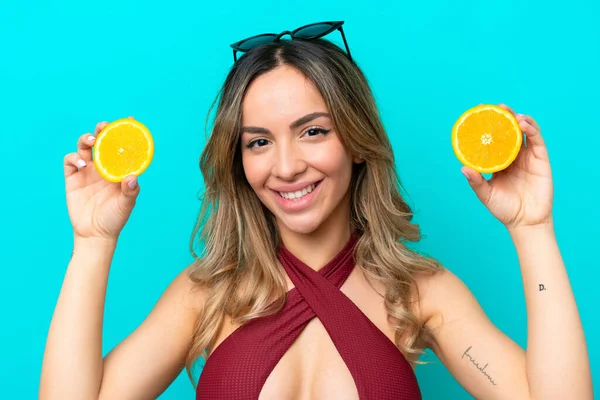 Mavi arka planda mayolu ve elinde portakal tutan beyaz bir kadın.