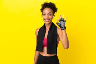 Sarı arka planda izole edilmiş genç Afrikalı spor kadını parmaklarıyla tamam işareti gösteriyor.