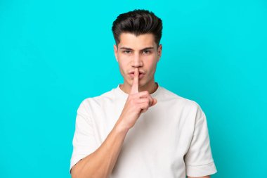 Genç, beyaz, mavi bakkalda izole edilmiş genç bir adam sessizlik işareti gösteriyor. Parmağını ağzına sok.