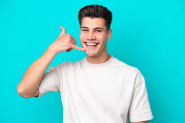 Genç, yakışıklı, beyaz bir adam, mavi bir bakkalda izole edilmiş, telefon jesti yapıyor. Bana geri dönüş işareti ver.