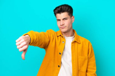 Genç, yakışıklı, beyaz bir adam, mavi bakkal bahçesinde izole edilmiş, olumsuz bir ifadeyle baş parmağını gösteriyor.