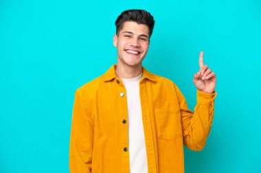 Genç, yakışıklı, beyaz bir adam, mavi bir bakkalda izole edilmiş ve en iyinin işareti olarak parmağını kaldırıyor.