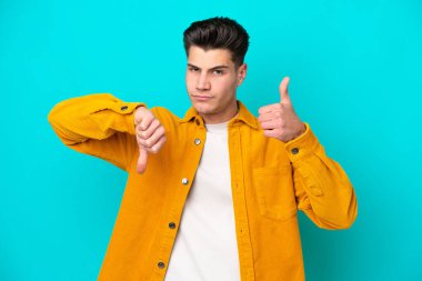 Young handsome caucasian man isolated on blue bakcground making good-bad sign. Undecided between yes or not