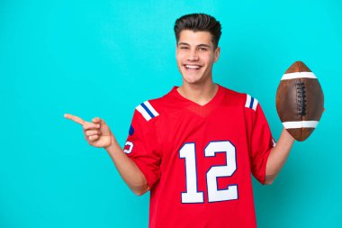 Genç beyaz adam rugby oynuyor. Mavi arka planda izole edilmiş. Parmağını yan tarafa gösteriyor.