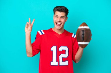 Genç beyaz adam rugby oynuyor. Mavi arka planda izole edilmiş. Gülümsüyor ve zafer işareti gösteriyor.