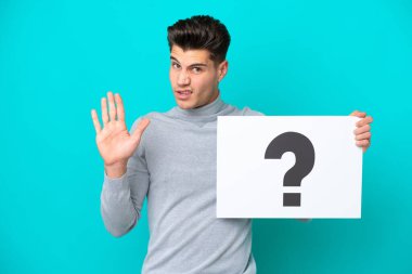 Mavi bakkalda izole edilmiş, üzerinde soru işareti olan bir pankart taşıyan yakışıklı beyaz bir adam.