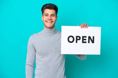 Mavi bakkalda izole edilmiş yakışıklı beyaz bir adam elinde mutlu bir ifadeyle açık bir pankart tutuyor.