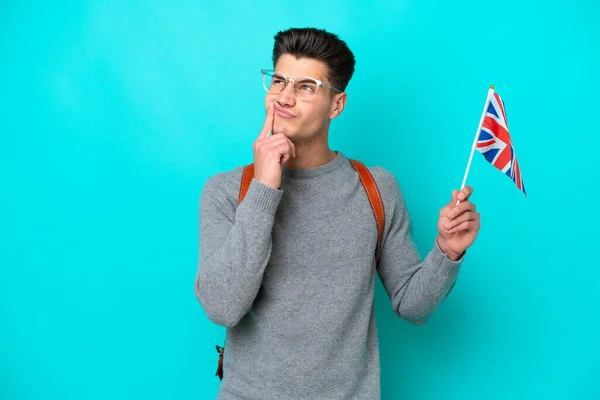年轻的高加索男子举着一面英国国旗 蓝色背景孤立 一边举目一边怀疑 — 图库照片