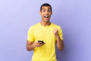 Zaferi kutlamak için cep telefonuyla izole edilmiş mor arka plan üzerinde Afrikalı Amerikalı bir adam.