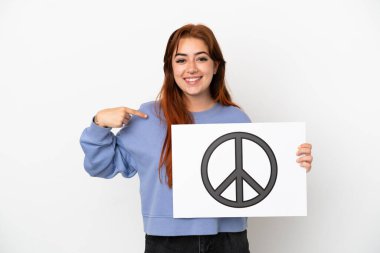 Genç kızıl saçlı kadın beyaz arka planda izole edilmiş. Elinde barış sembolü olan bir pankart var ve onu işaret ediyor.