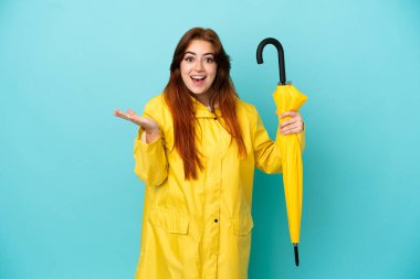 Elinde şemsiyesi olan kızıl saçlı kadın mavi arka planda izole edilmiş ve şok olmuş bir yüz ifadesi var.