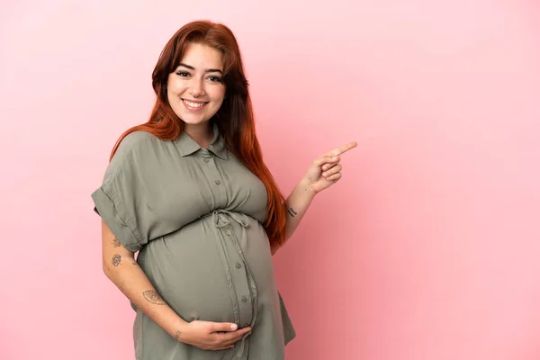 妊娠中のピンクの背景に隔離され 横を指している若い赤毛の白人女性 — ストック写真