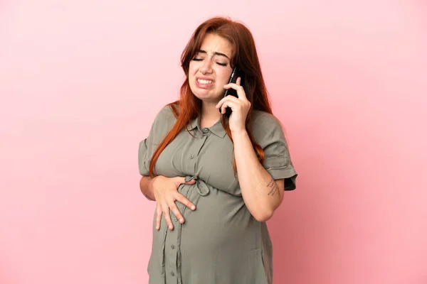 妊娠中や携帯電話を使用してピンクの背景に隔離された若い赤毛の白人女性 — ストック写真