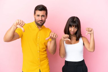 Pembe arka planda izole edilmiş genç bir çift baş parmağını iki eliyle aşağı gösteriyor.