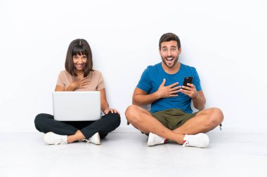 Yerde oturan genç çift beyaz arka planda izole edilmiş cep telefonu ve bilgisayarla ellerini göğsüne koyup gülümsüyor.
