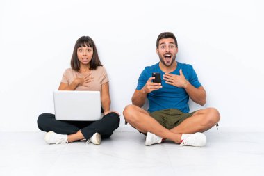 Yerde oturan genç çift beyaz arka planda izole edilmiş bilgisayar ve cep telefonlarını tutarken şaşırmış ve şok olmuştu.