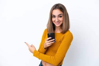 Beyaz arka planda izole edilmiş genç beyaz bir kadın cep telefonu kullanıyor ve geriye doğru işaret ediyor.