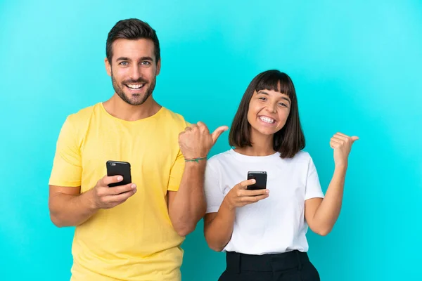 Casal Jovem Isolado Fundo Azul Usando Telefone Celular Apontando Para — Fotografia de Stock