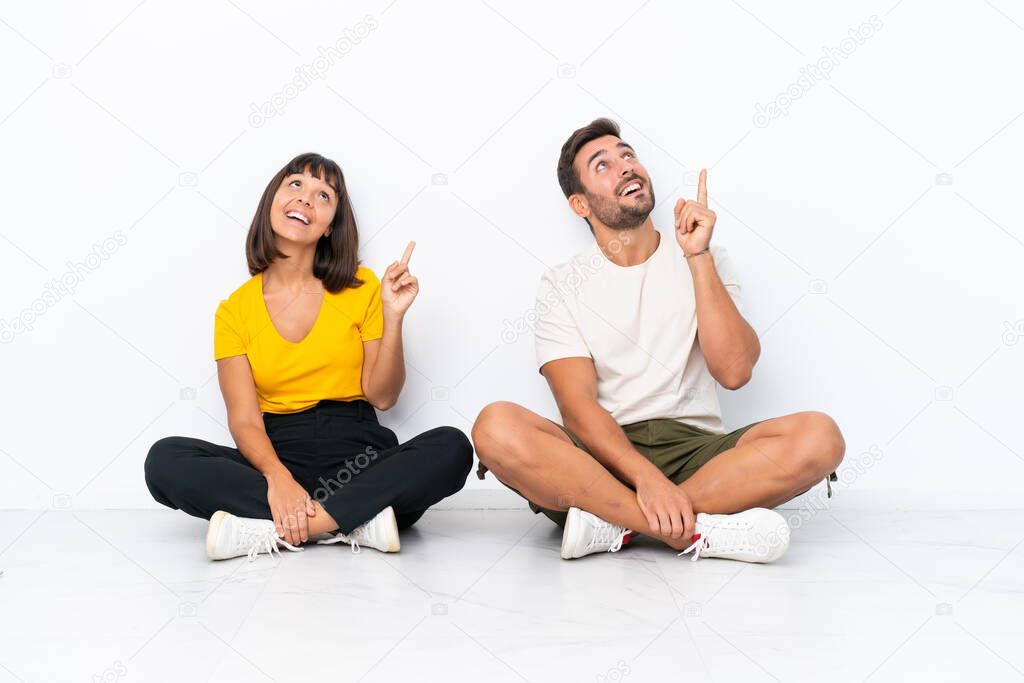 Young couple sitting on the floor isolated on white background pointing a great idea and looking up
