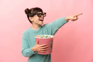 Küçük beyaz kız pembe arka planda 3 boyutlu gözlüklerle izole edilmiş ve elinde koca bir kova patlamış mısırla...