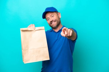 Orta yaşlı bir adam mavi arka planda izole edilmiş yiyecek paketini alıp mutlu bir ifadeyle cepheyi işaret ediyor.