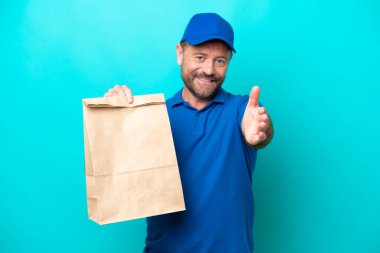 Orta yaşlı bir adam mavi arka planda izole edilmiş bir paket yiyecek alıyor ve iyi bir anlaşma için el sıkışıyor.