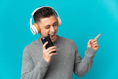 Kafkasyalı genç adam mavi arka planda izole edilmiş. Cep telefonuyla müzik dinliyor ve şarkı söylüyor.