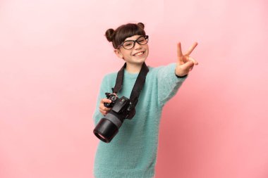 Küçük fotoğrafçı kız pembe arka planda izole edilmiş gülümsüyor ve zafer işareti gösteriyor.