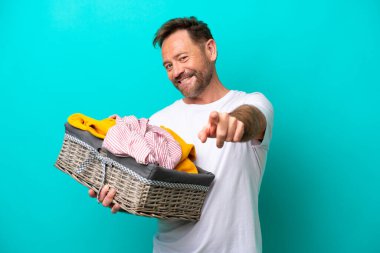 Mavi arka planda izole edilmiş çamaşır sepetini tutan orta yaşlı kadın mutlu bir ifadeyle ön tarafı işaret ediyor.